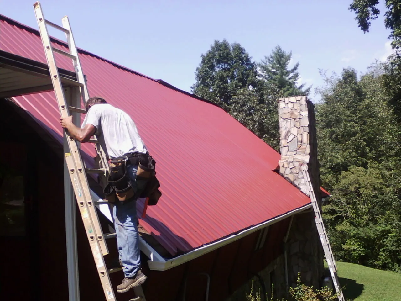Roofer on a ladder beside a red metal roof and stone chimney — Soffit & Fascia Repair by Roofing Brothers in Elrama